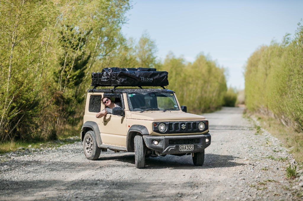 Suzuki Jimny on a New Zealand gravel road with rooftop tent, customer waving from the driver's window